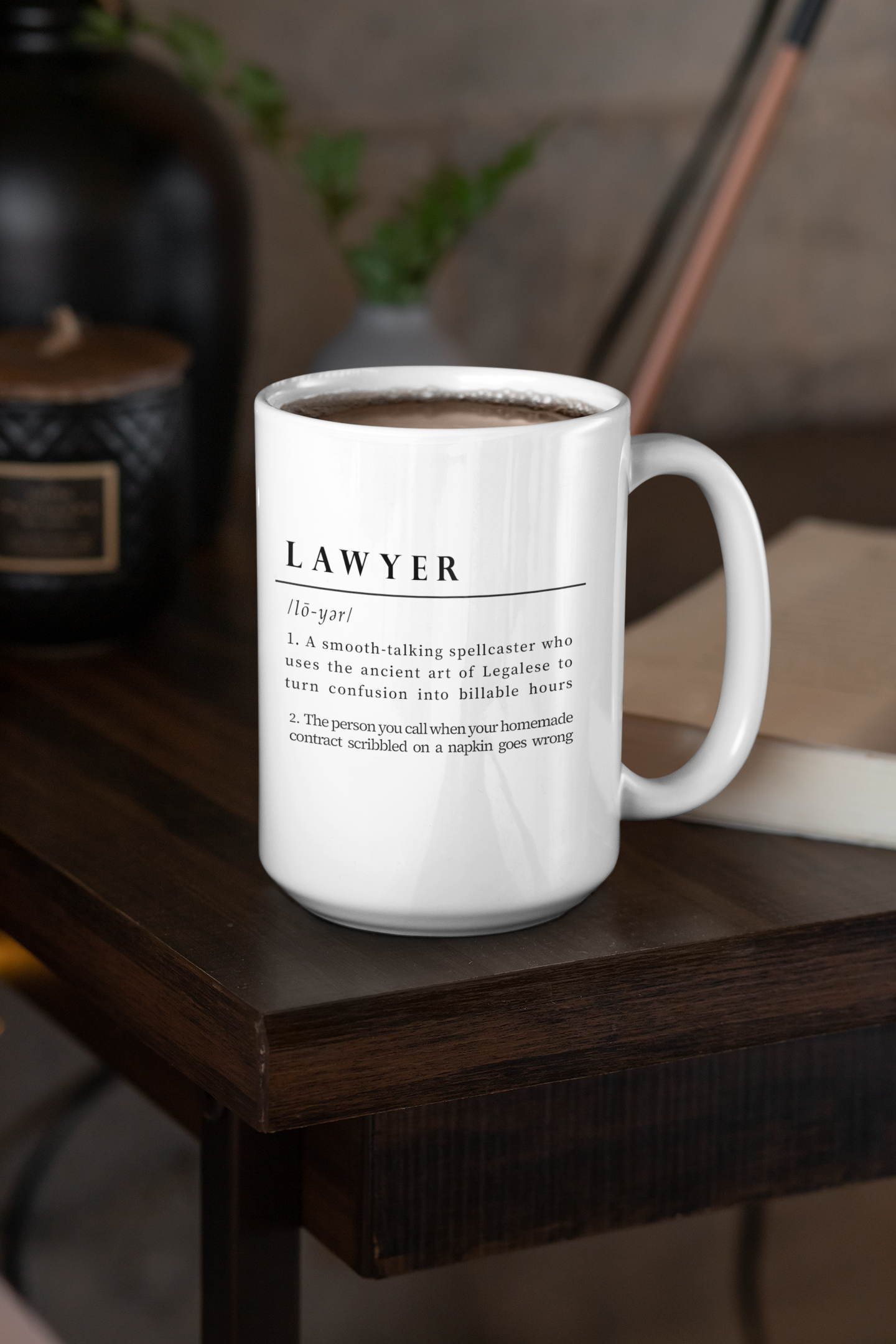 A white ceramic mug displays a playful and witty definition of 'Lawyer', printed in a bold black font. The text humorously depicts a lawyer as a spellcaster in Legalese and a savior of napkin contracts. 