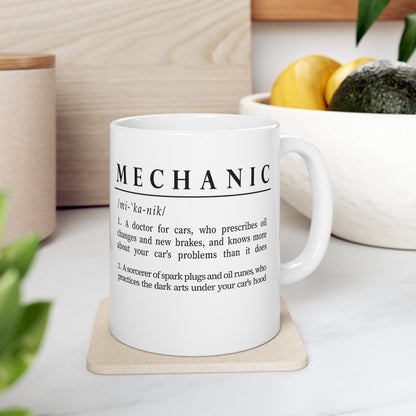 Displayed is a white ceramic mug with a witty definition of 'Mechanic' in bold black font. The text creatively describes a mechanic as both a car doctor and a sorcerer of automotive parts, adding a humorous and mythical element to the profession. 