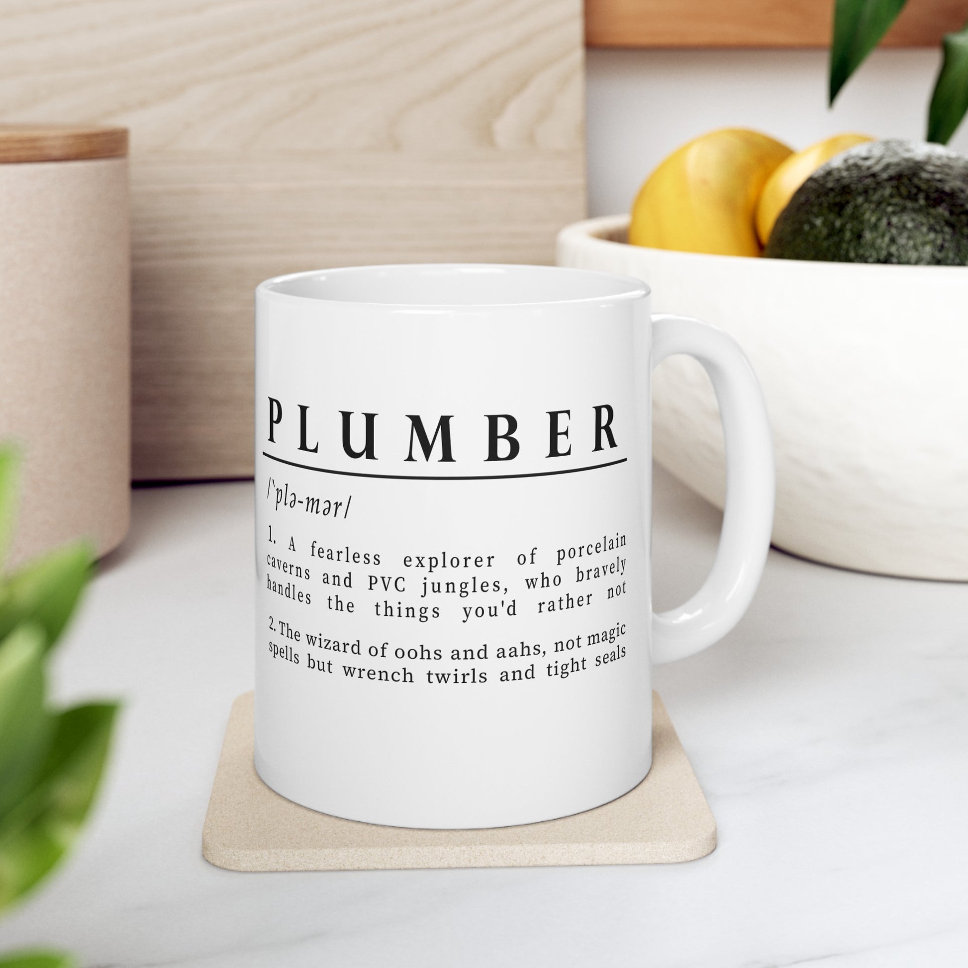 white ceramic mug with a witty definition of 'Plumber' printed in black. The humorous description highlights a plumber as both an explorer of pipes and a skilled artisan.