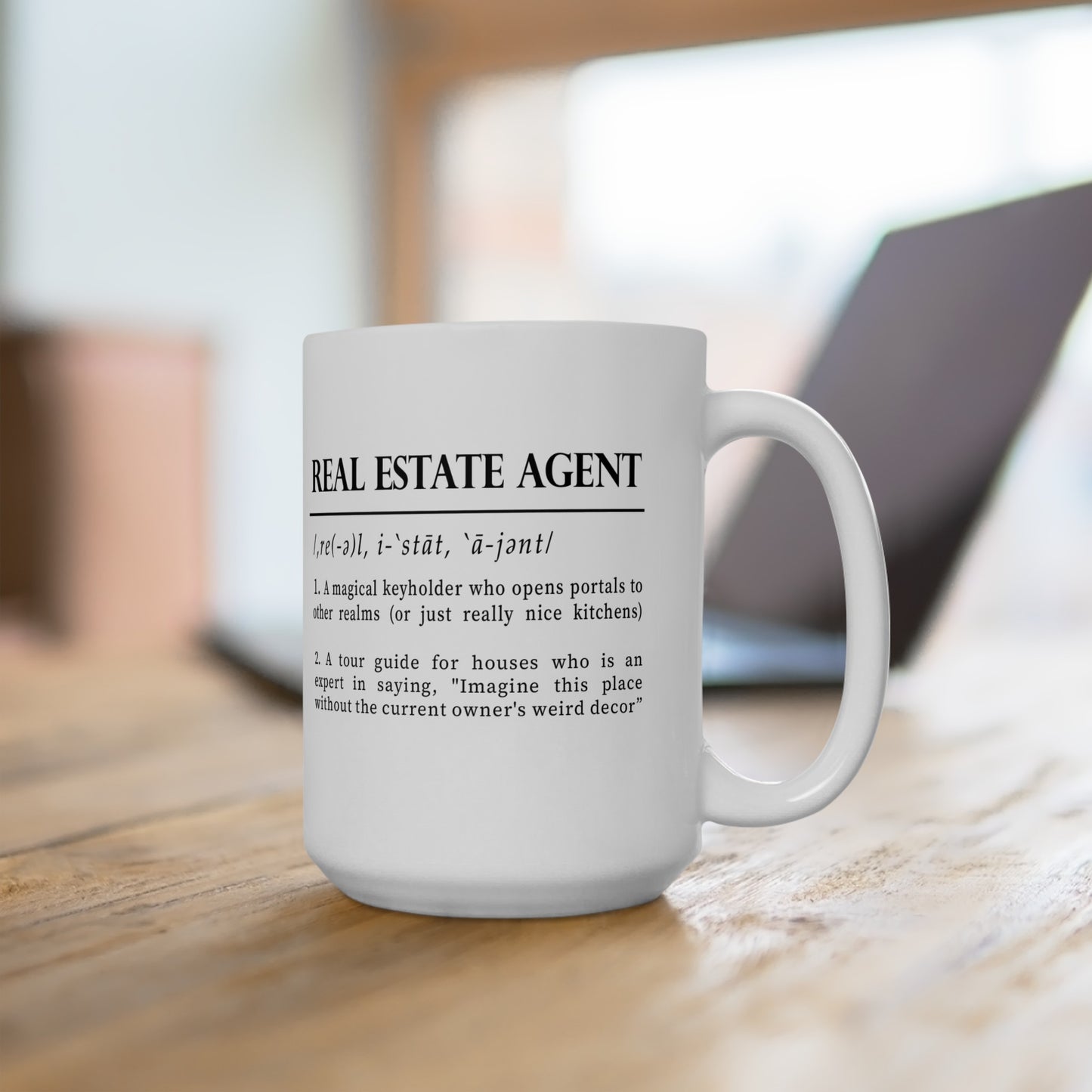 A classic white ceramic mug features a playful definition of 'Real Estate Agent' printed in elegant black font. The text humorously describes the agent as a keyholder to realms and an expert in home decor imagination. 