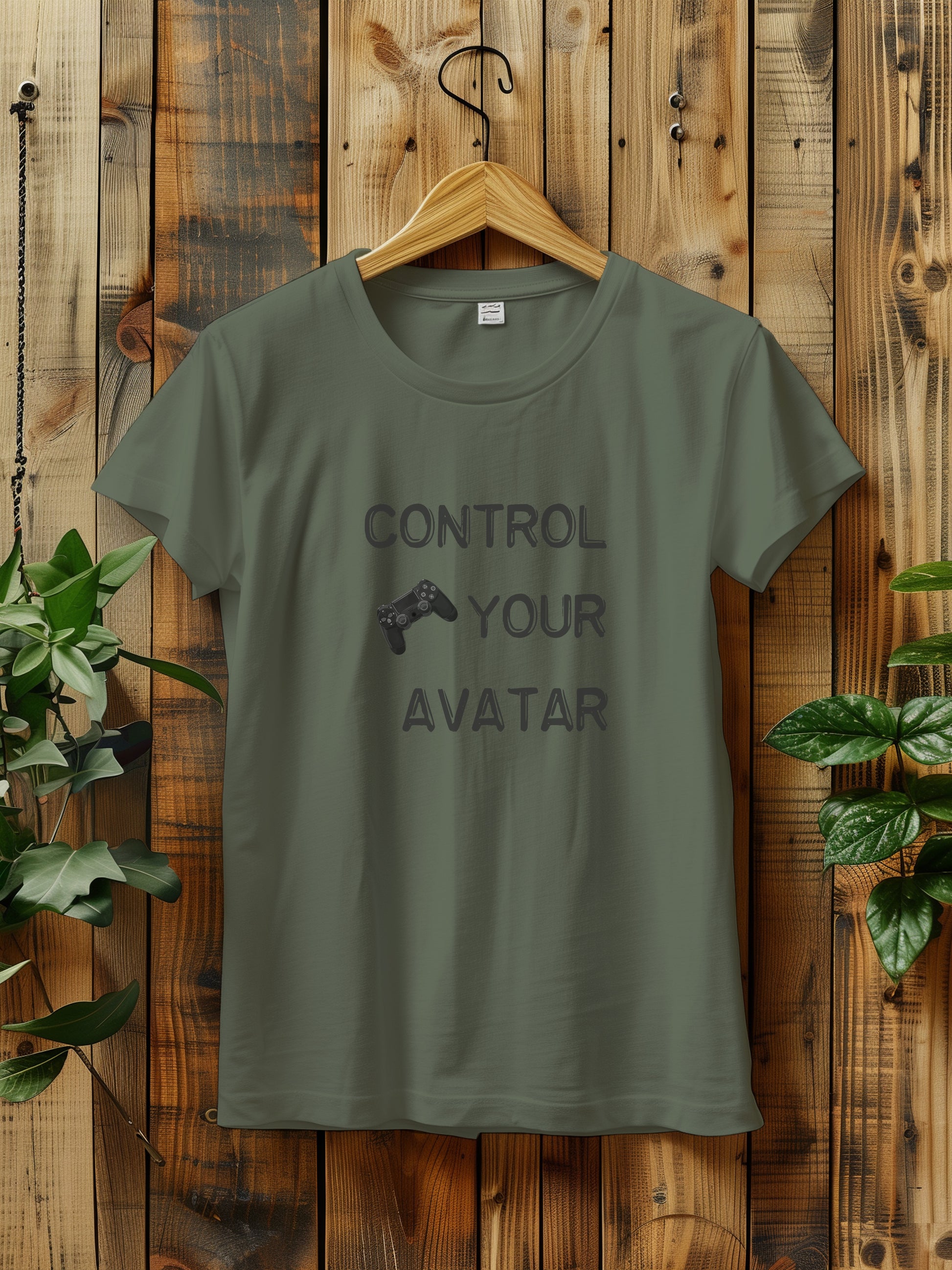 T-shirt with the text "CONTROL YOUR AVATAR" and an illustration of a game controller, hanging on a wooden wall. The scene is decorated with green plants on either side, creating a rustic and natural vibe