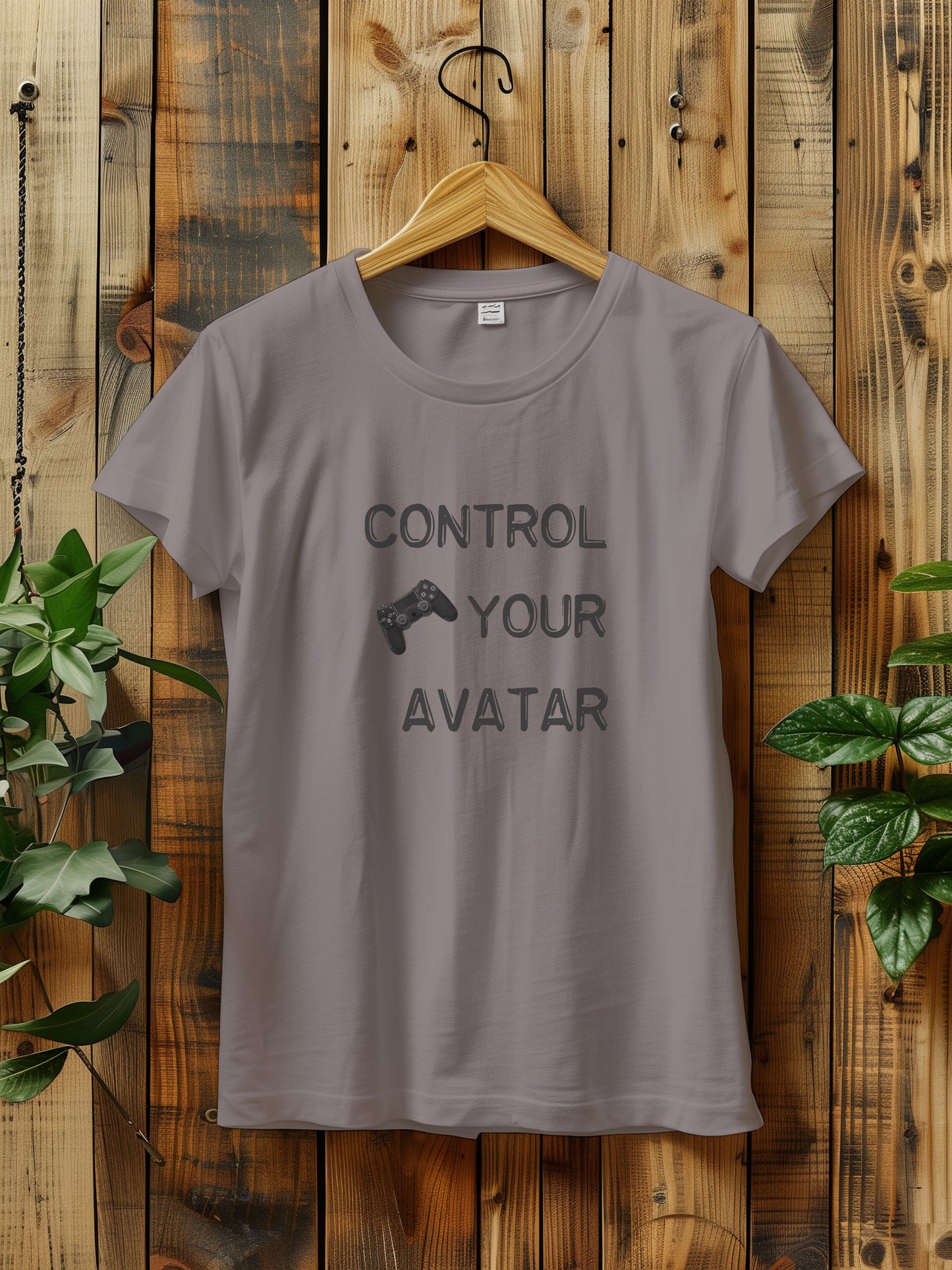 T-shirt with the text "CONTROL YOUR AVATAR" and an illustration of a game controller, hanging on a wooden wall. The scene is decorated with green plants on either side, creating a rustic and natural vibe