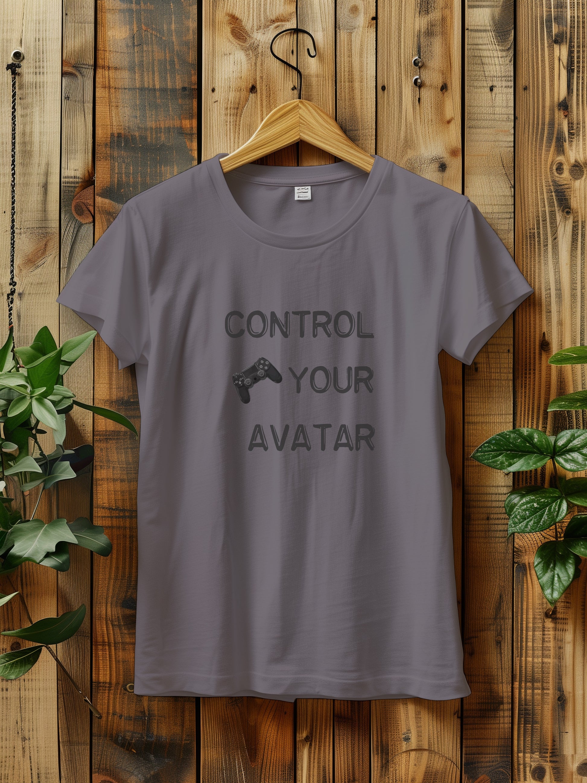 T-shirt with the text "CONTROL YOUR AVATAR" and an illustration of a game controller, hanging on a wooden wall. The scene is decorated with green plants on either side, creating a rustic and natural vibe