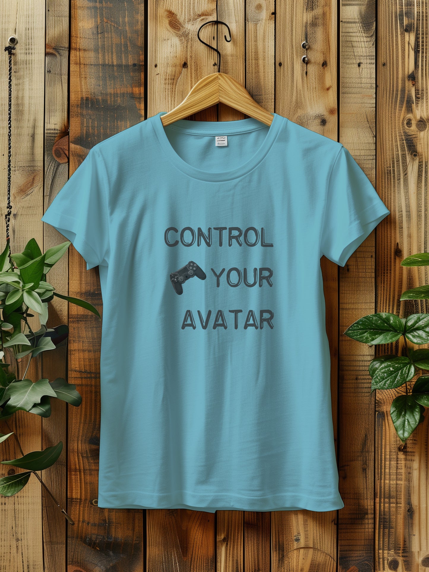 T-shirt with the text "CONTROL YOUR AVATAR" and an illustration of a game controller, hanging on a wooden wall. The scene is decorated with green plants on either side, creating a rustic and natural vibe