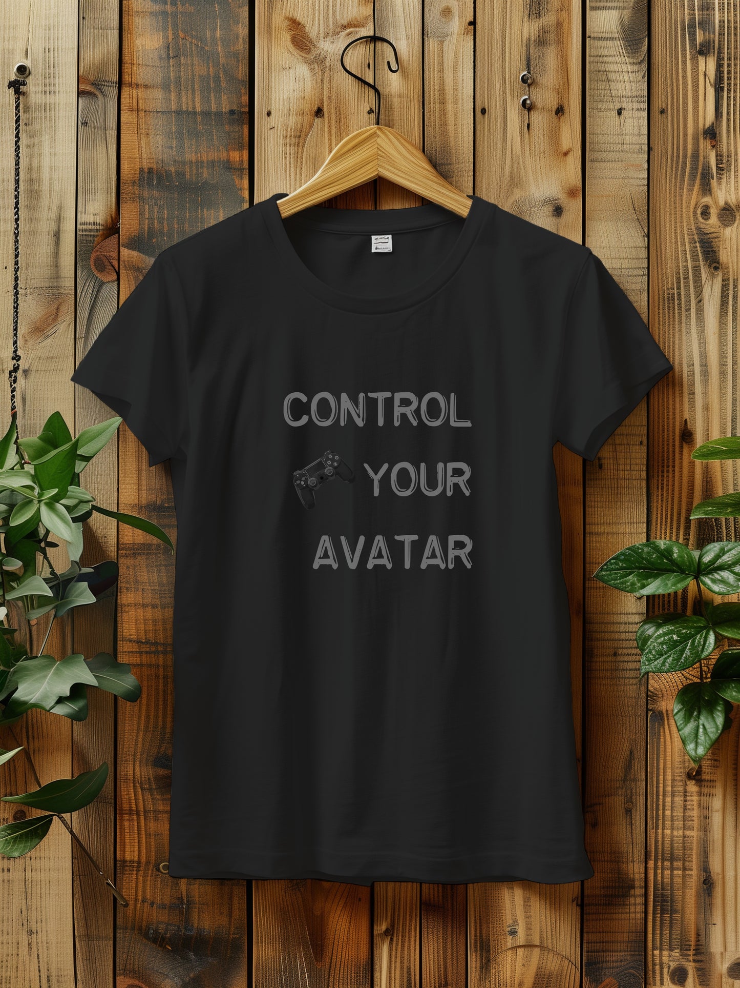 T-shirt with the text "CONTROL YOUR AVATAR" and an illustration of a game controller, hanging on a wooden wall. The scene is decorated with green plants on either side, creating a rustic and natural vibe