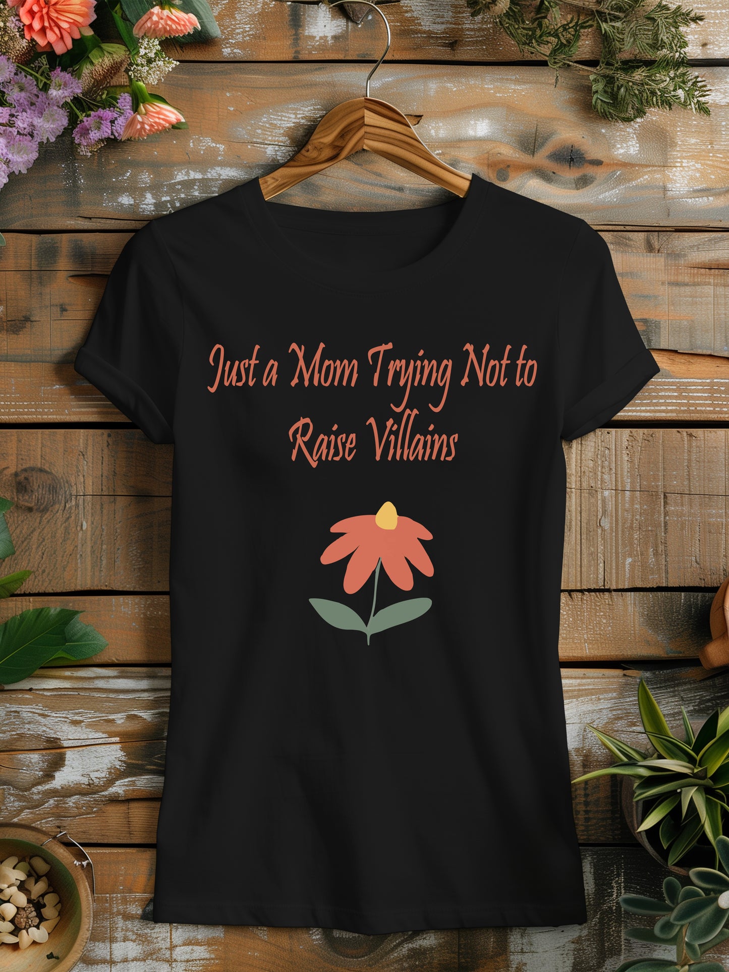 t-shirt featuring the humorous phrase "Just a Mom Trying Not to Raise Villains" in script, accompanied by a simple orange and green flower illustration.