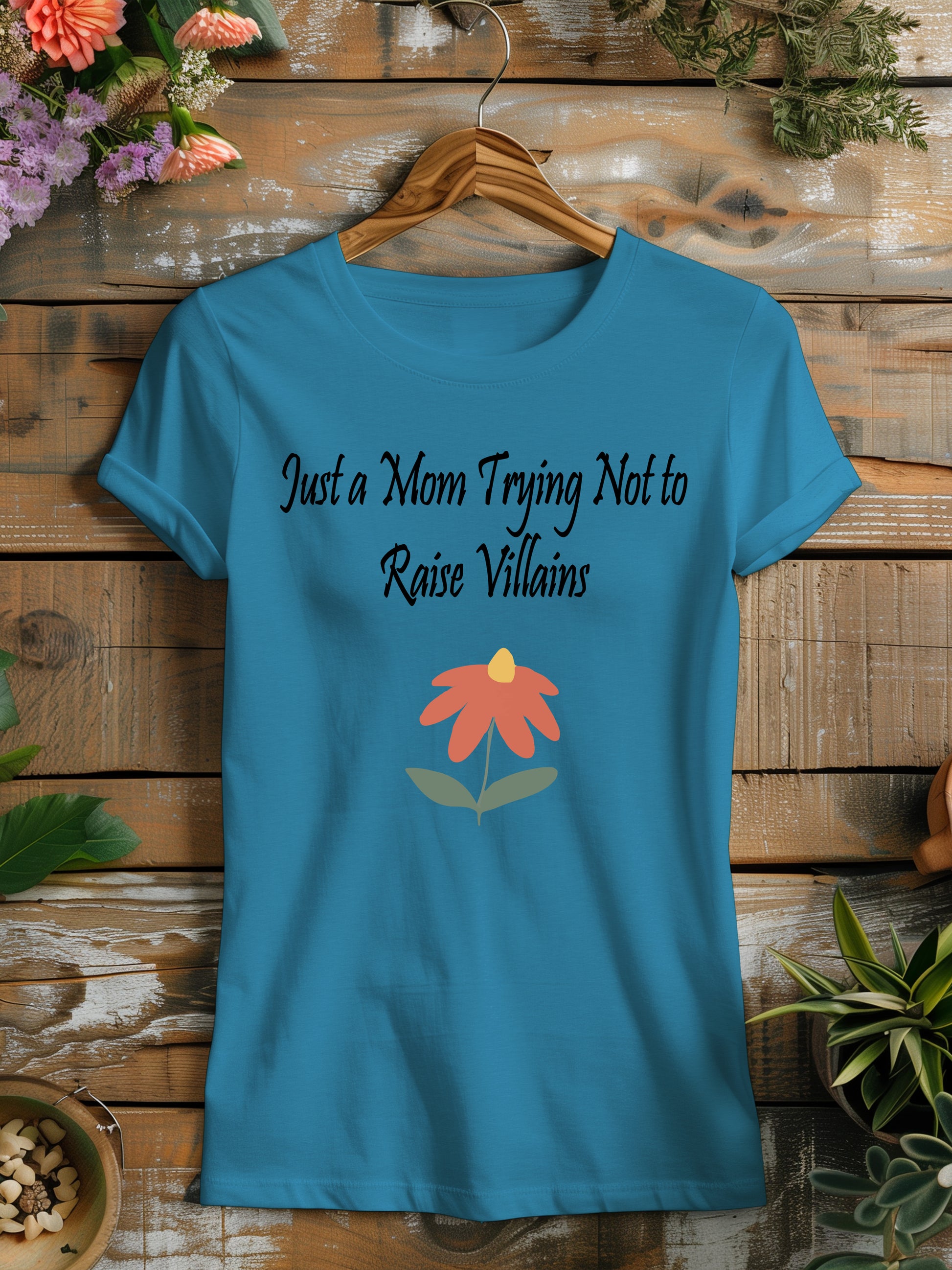 t-shirt featuring the humorous phrase "Just a Mom Trying Not to Raise Villains" in black script, accompanied by a simple orange and green flower illustration.