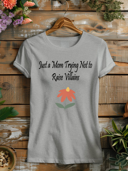 t-shirt featuring the humorous phrase "Just a Mom Trying Not to Raise Villains" in black script, accompanied by a simple orange and green flower illustration.