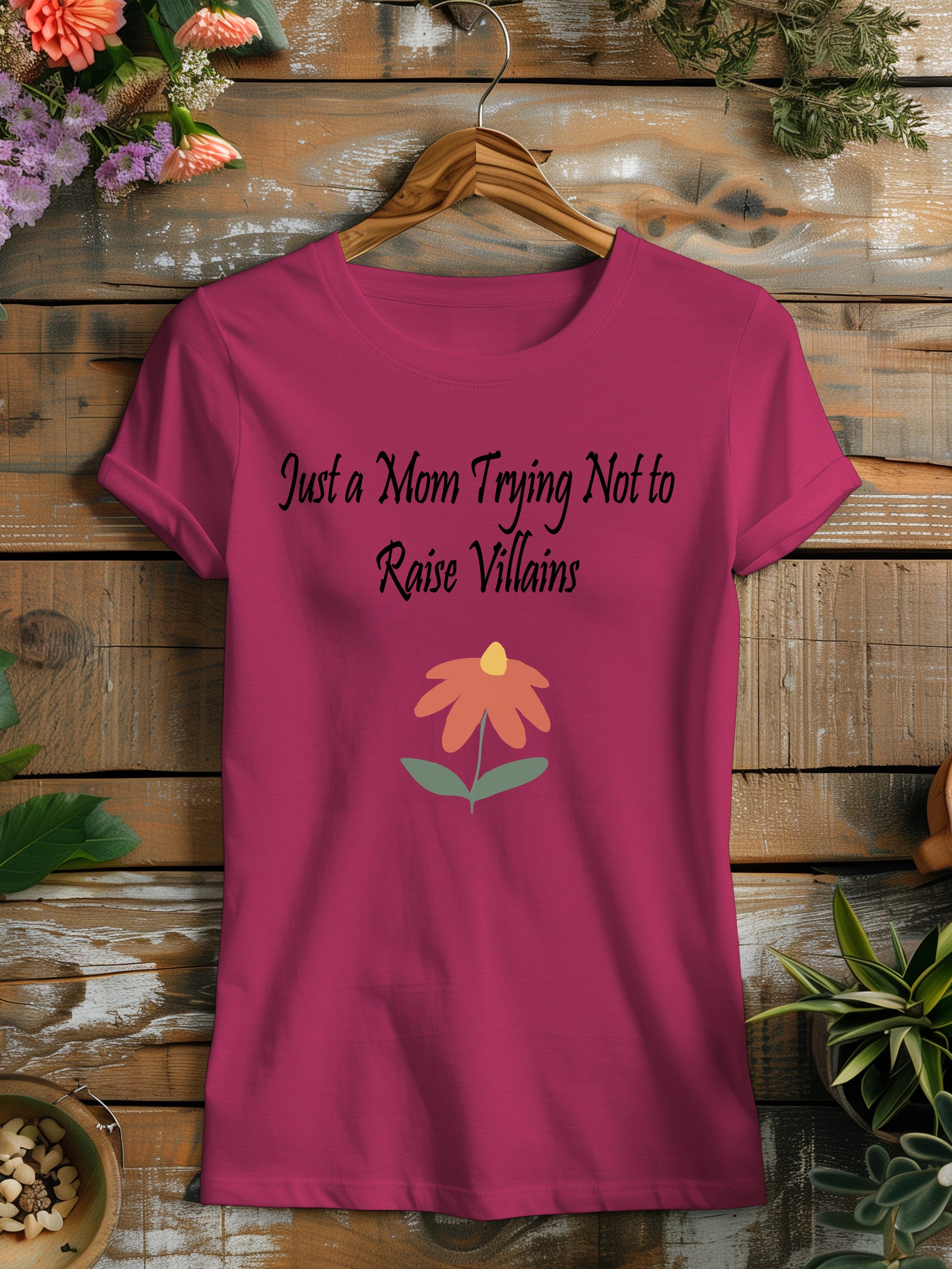 t-shirt featuring the humorous phrase "Just a Mom Trying Not to Raise Villains" in black script, accompanied by a simple orange and green flower illustration.