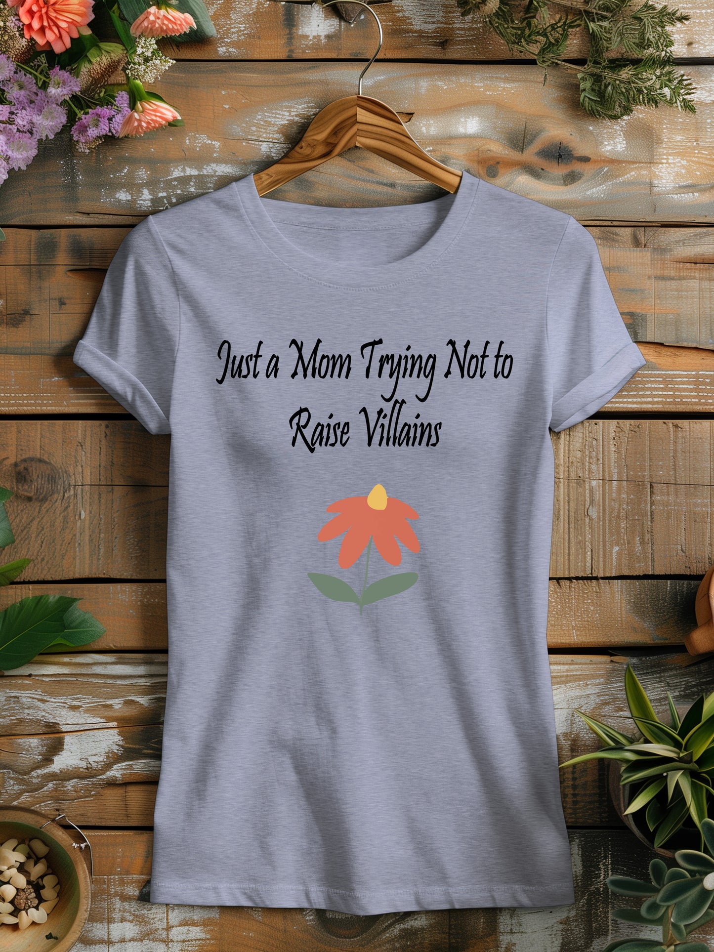 t-shirt featuring the humorous phrase "Just a Mom Trying Not to Raise Villains" in black script, accompanied by a simple orange and green flower illustration.