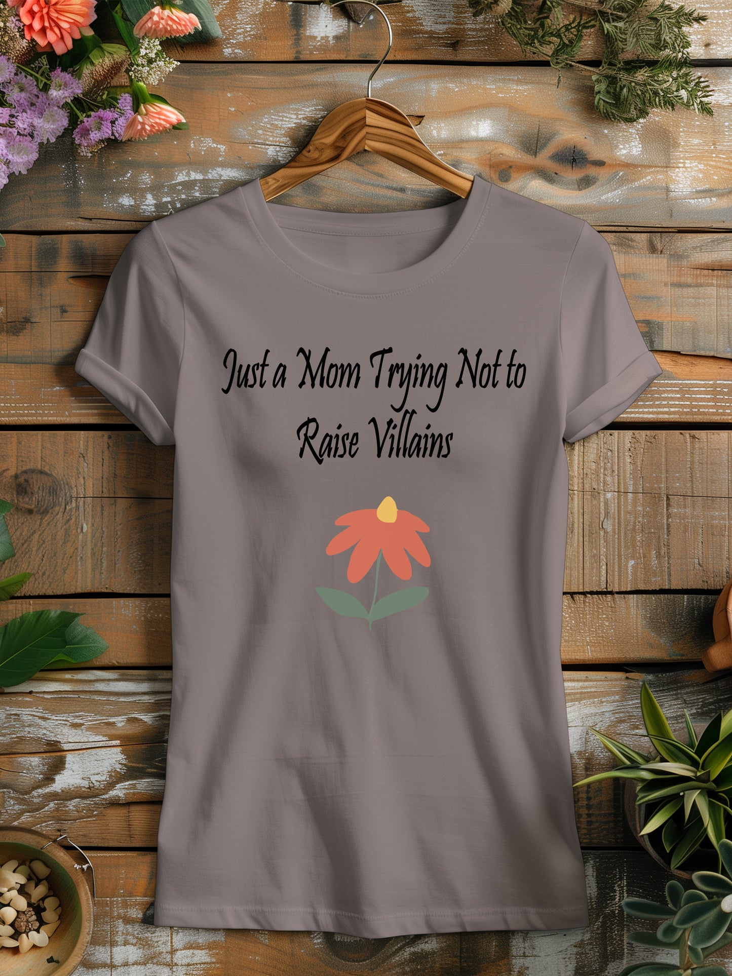 t-shirt featuring the humorous phrase "Just a Mom Trying Not to Raise Villains" in black script, accompanied by a simple orange and green flower illustration.