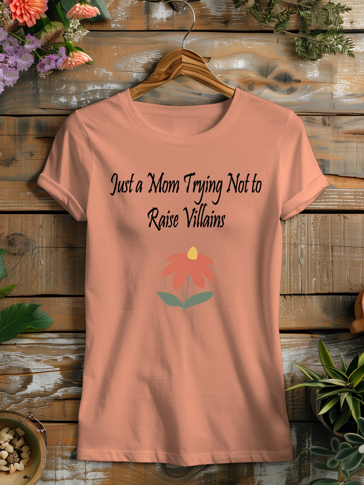 t-shirt featuring the humorous phrase "Just a Mom Trying Not to Raise Villains" in black script, accompanied by a simple orange and green flower illustration.