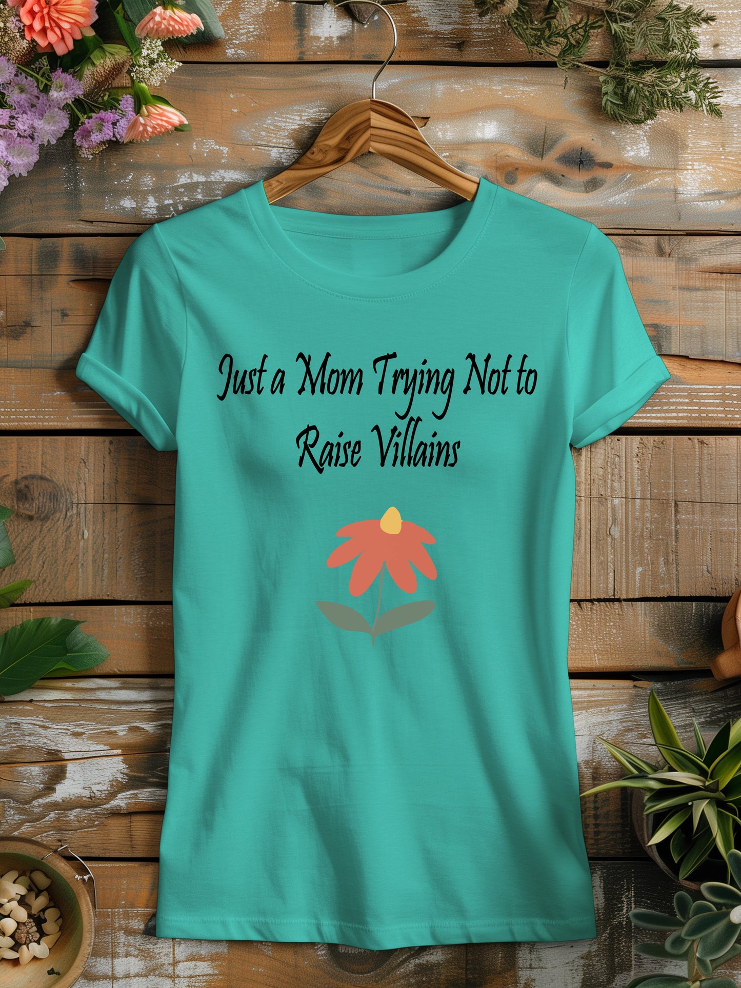 t-shirt featuring the humorous phrase "Just a Mom Trying Not to Raise Villains" in black script, accompanied by a simple orange and green flower illustration.