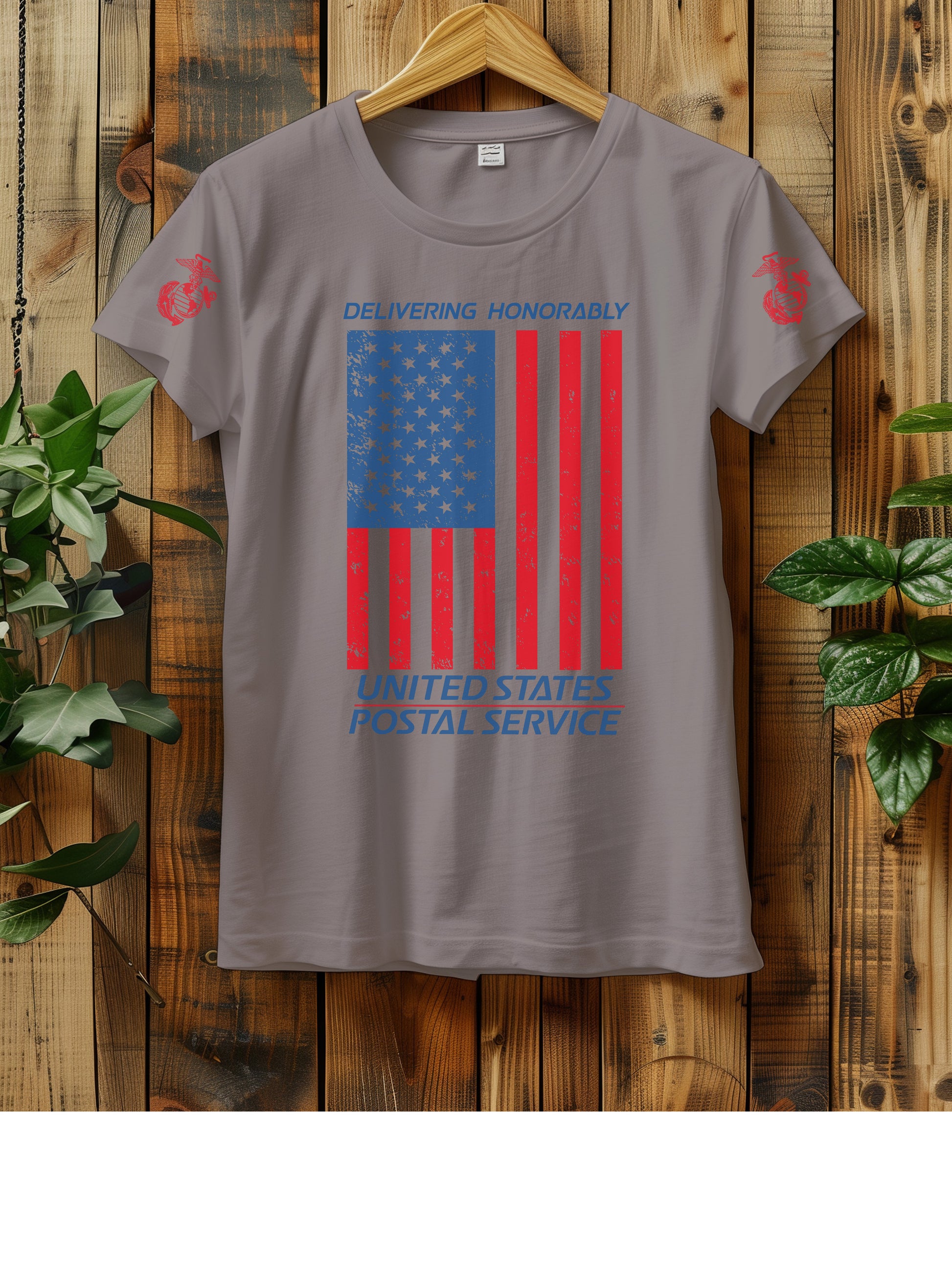 T-shirt hanging on a wooden wall with a hanger, displaying a distressed American flag graphic. The text "Delivering Honorably United States Postal Service" is printed in blue and red. The left sleeve has a red Marine Corps emblem. Two green plants are visible on either side of the shirt.