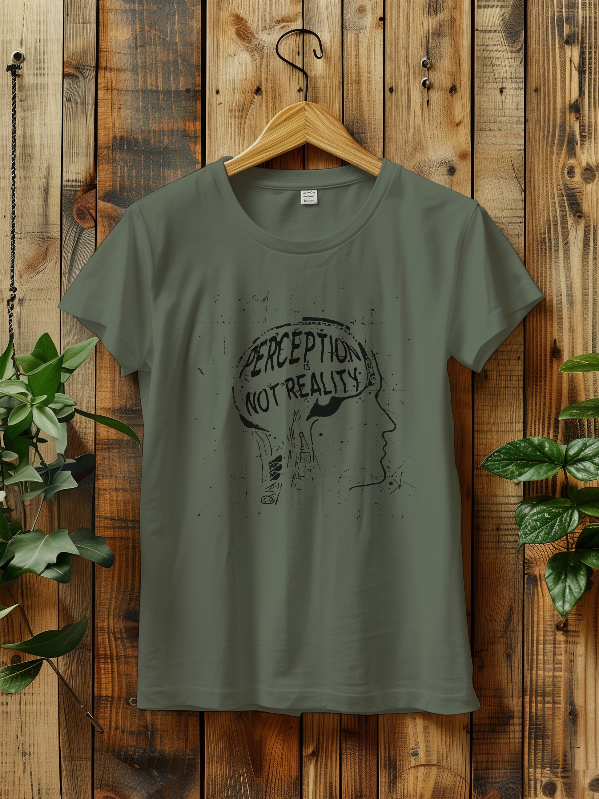 t-shirt featuring a graphic with the phrase "Perception is Not Reality" and a sketch of a head, hanging on a rustic wooden wall. The background includes green plants adding a natural touch to the scene.