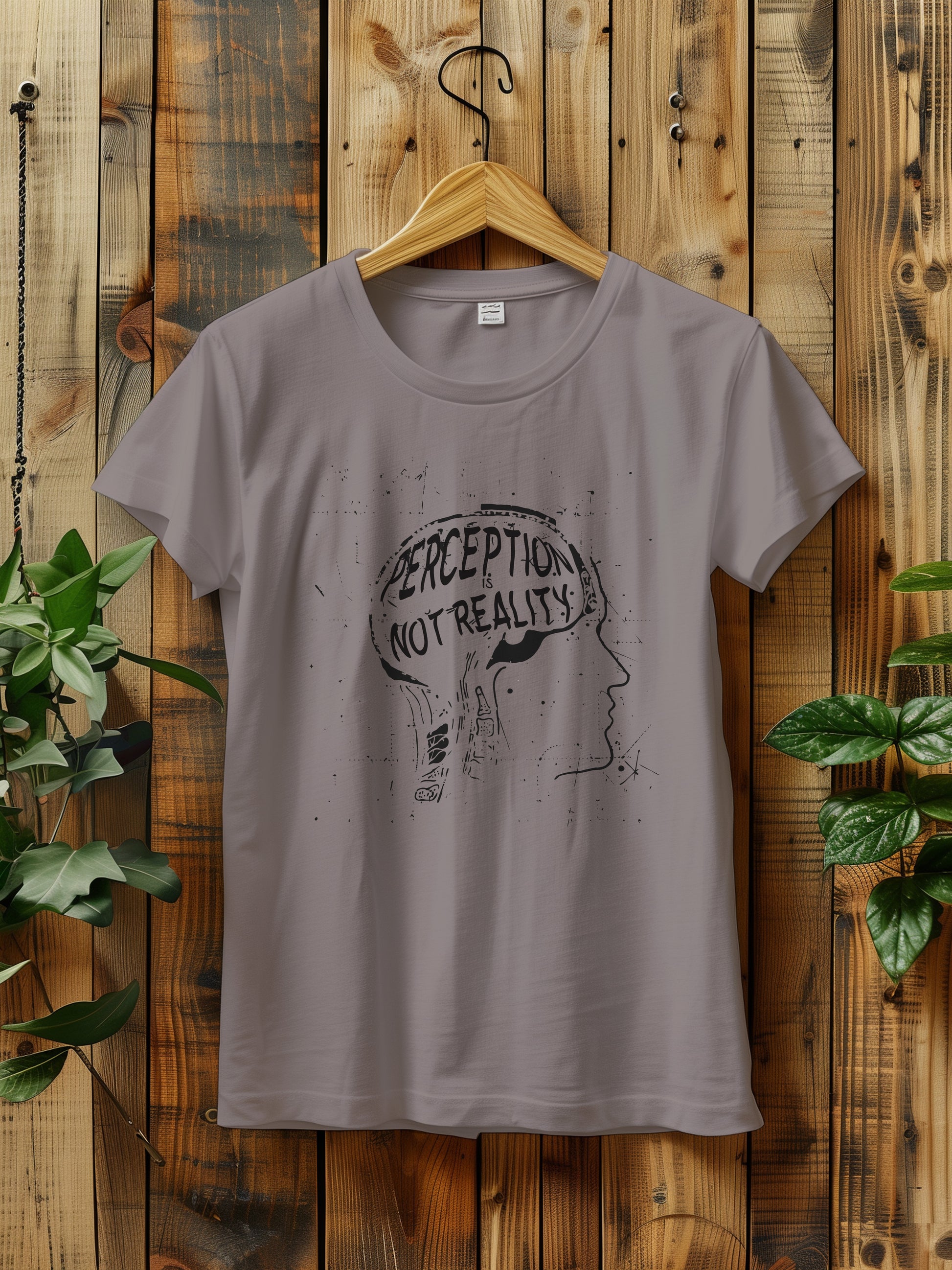 t-shirt featuring a graphic with the phrase "Perception is Not Reality" and a sketch of a head, hanging on a rustic wooden wall. The background includes green plants adding a natural touch to the scene.