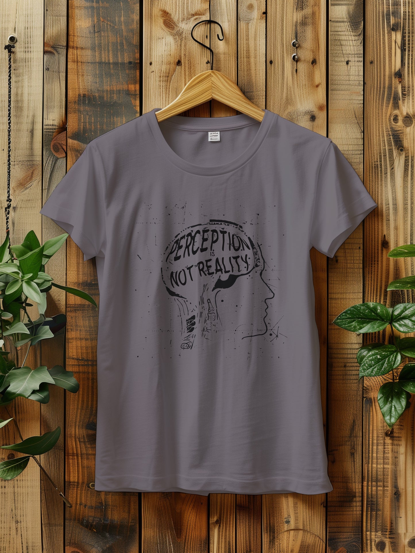 t-shirt featuring a graphic with the phrase "Perception is Not Reality" and a sketch of a head, hanging on a rustic wooden wall. The background includes green plants adding a natural touch to the scene.