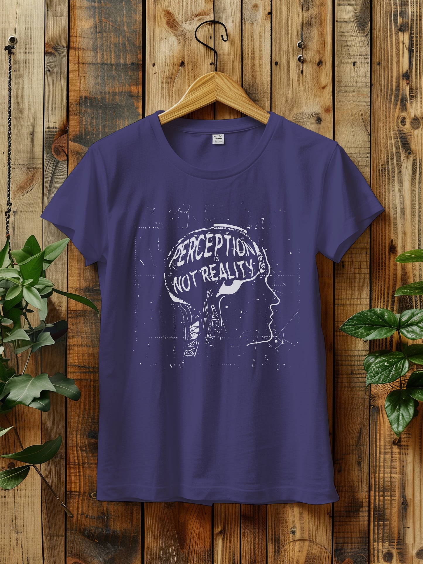 t-shirt featuring a graphic with the phrase "Perception is Not Reality" and a sketch of a head, hanging on a rustic wooden wall. The background includes green plants adding a natural touch to the scene.