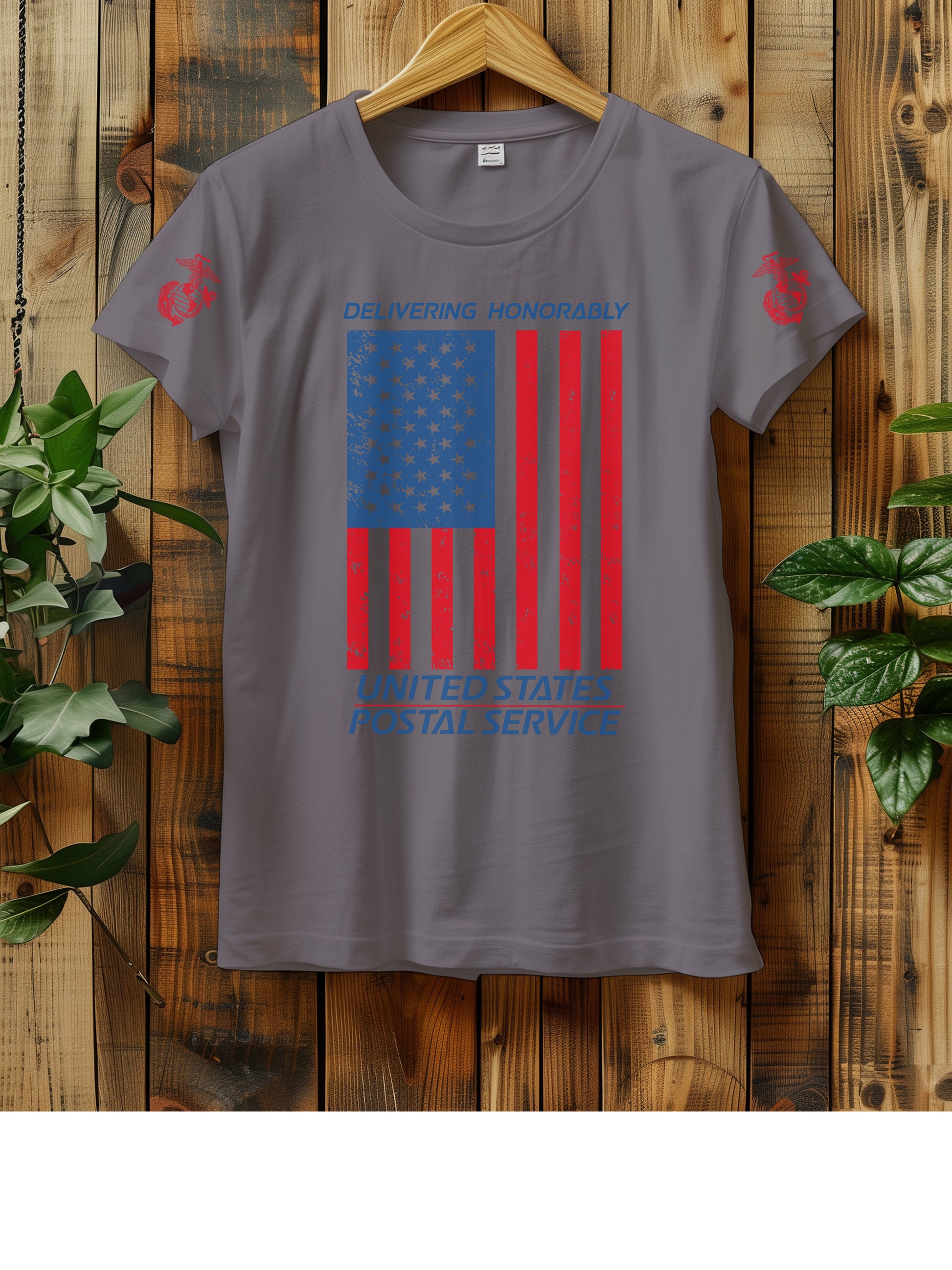 T-shirt hanging on a wooden wall with a hanger, displaying a distressed American flag graphic. The text "Delivering Honorably United States Postal Service" is printed in blue and red. The left sleeve has a red Marine Corps emblem. Two green plants are visible on either side of the shirt.