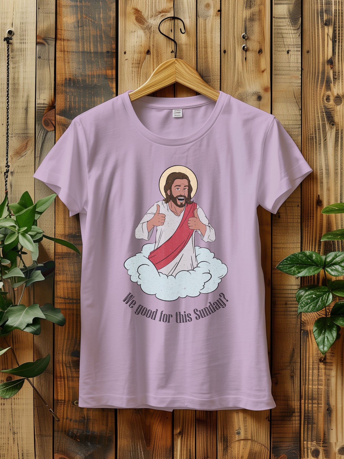 t-shirt with a cartoon of Jesus giving thumbs up, hanging on a wooden hanger against a wooden background.