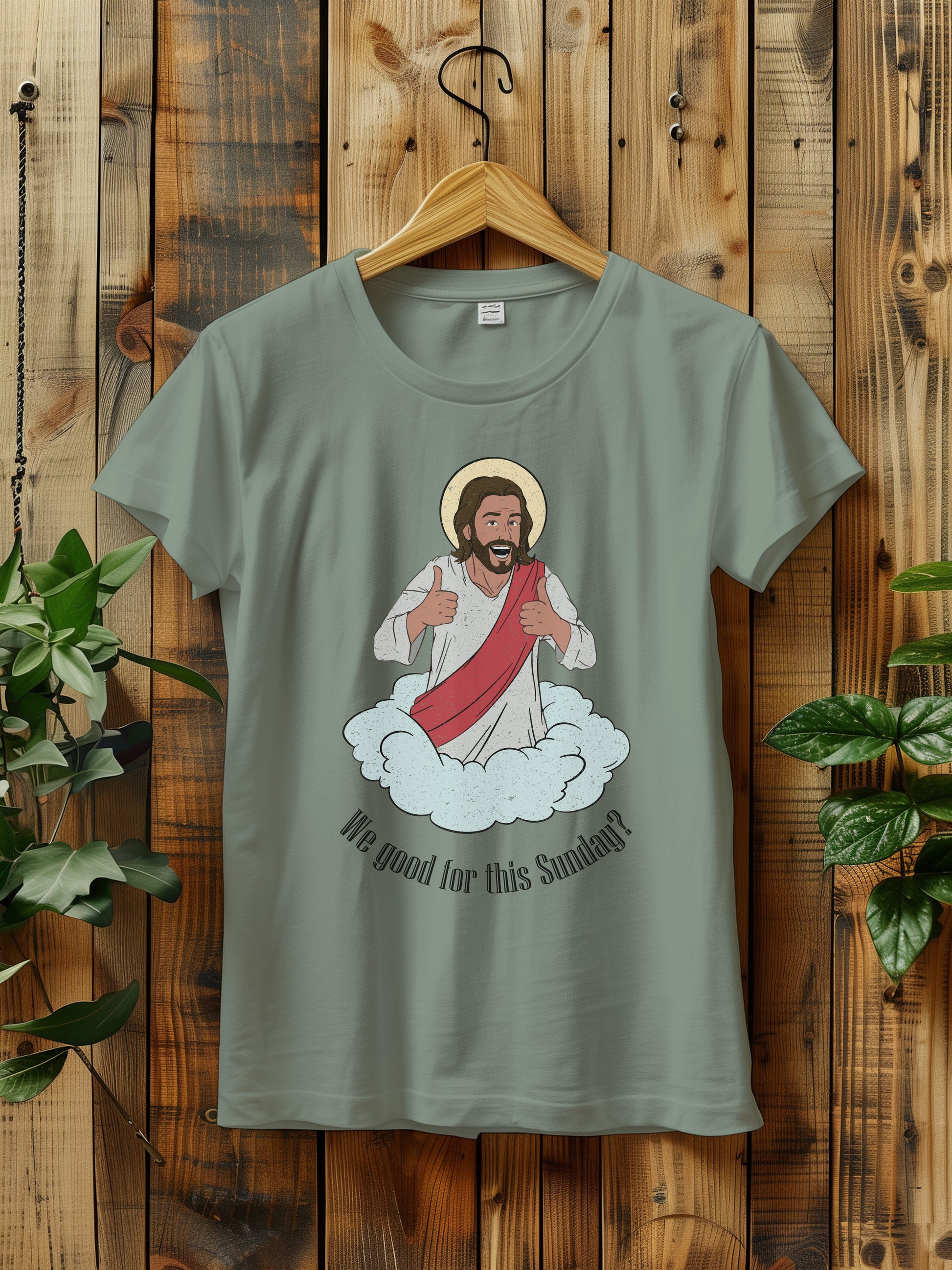 t-shirt with a cartoon of Jesus giving thumbs up, hanging on a wooden hanger against a wooden background.