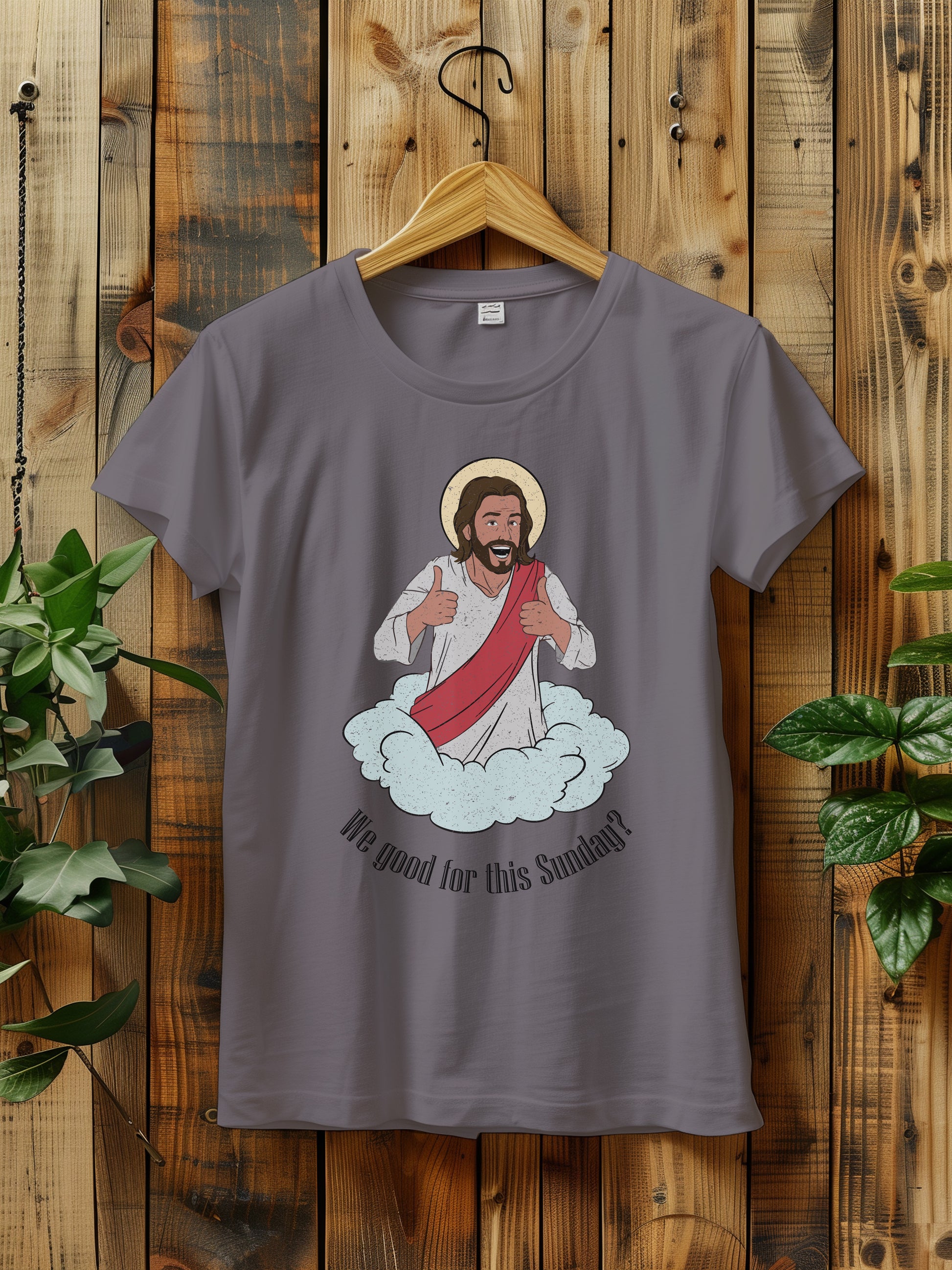 t-shirt with a cartoon of Jesus giving thumbs up, hanging on a wooden hanger against a wooden background.