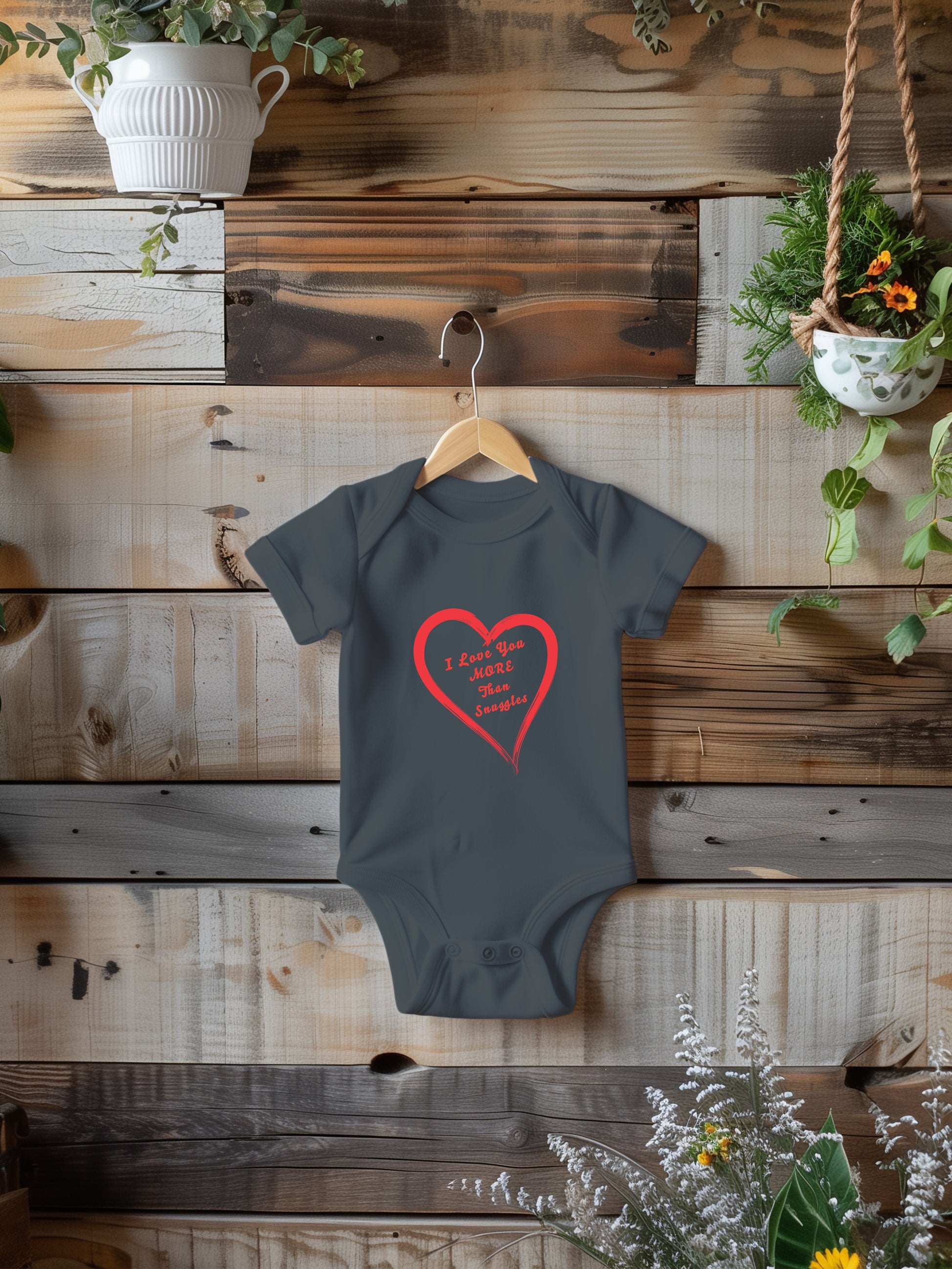 onesie with "I love you more than snuggles" in a heart on a wooden background with plants.